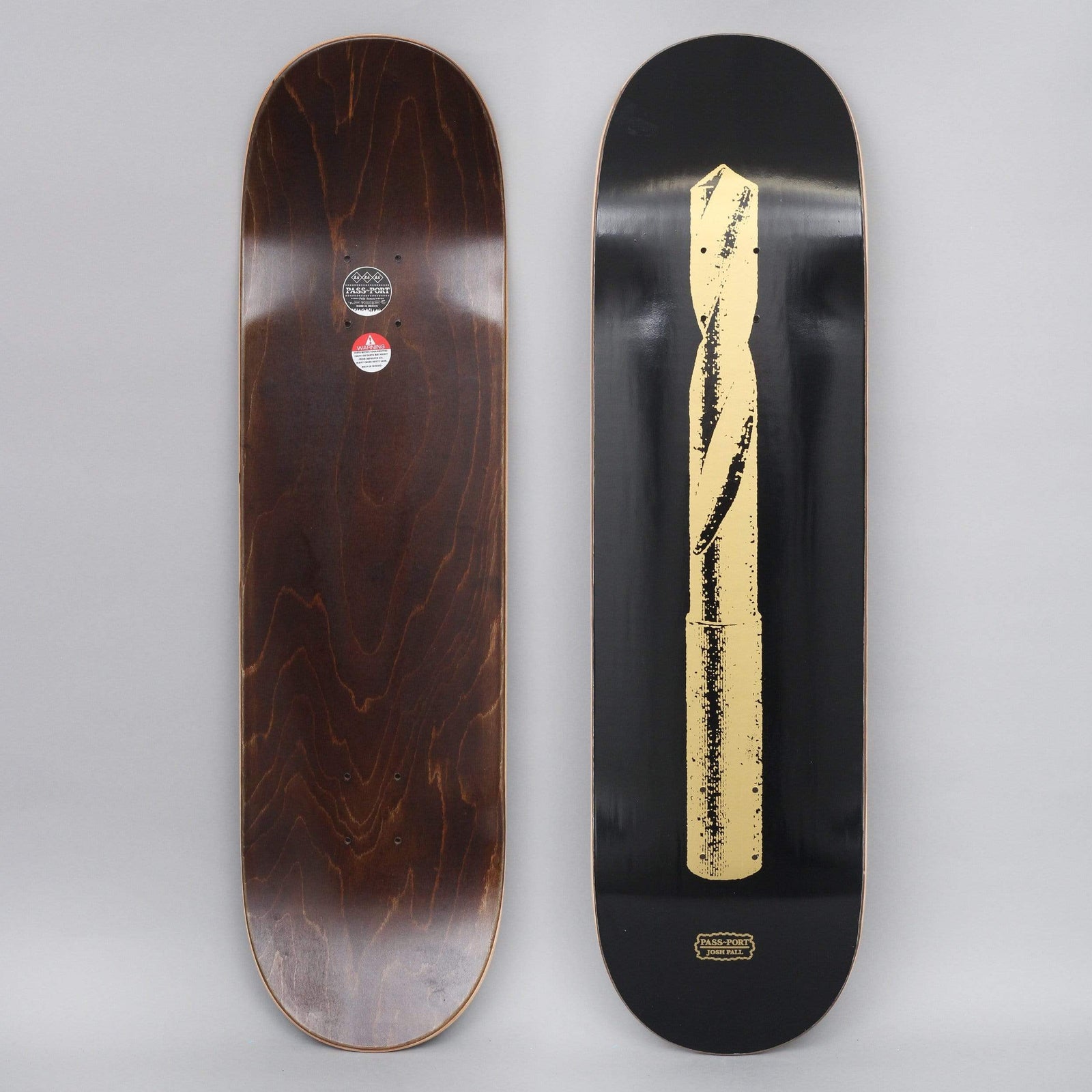 Passport Josh Pall Pro Series Black skateboard deck: A single large gold-coloured twist drill bit, printed in a rough, worn style, running the length of a black deck. A small Pass-Port logo with the rider's name sits at the tail.