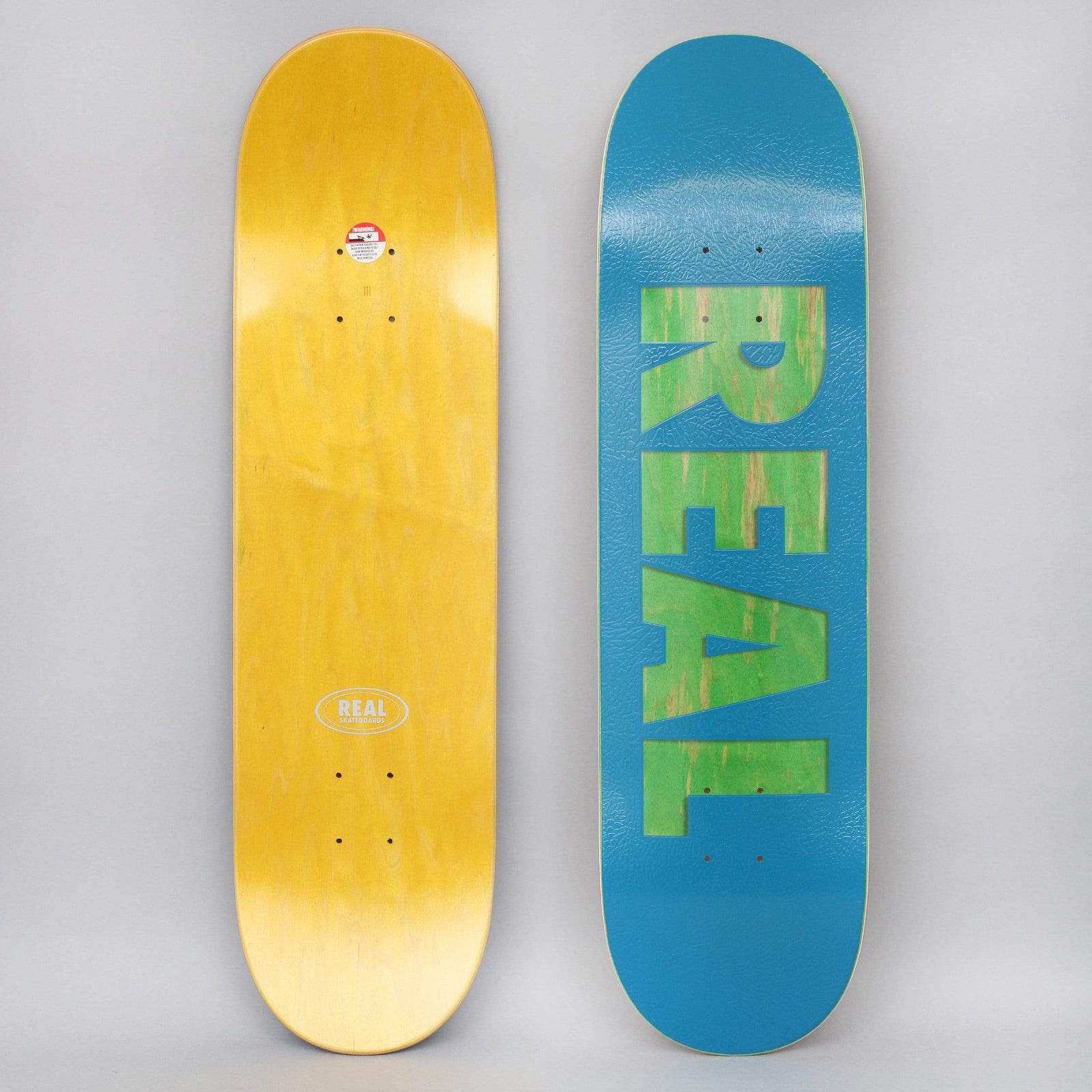 Real Bold Series Blue skateboard deck: Blue deck with the REAL name in large bold block capitals cut through to reveal a green wood-grain ply. Yellow-stained underside with the Real oval logo shown beside it.