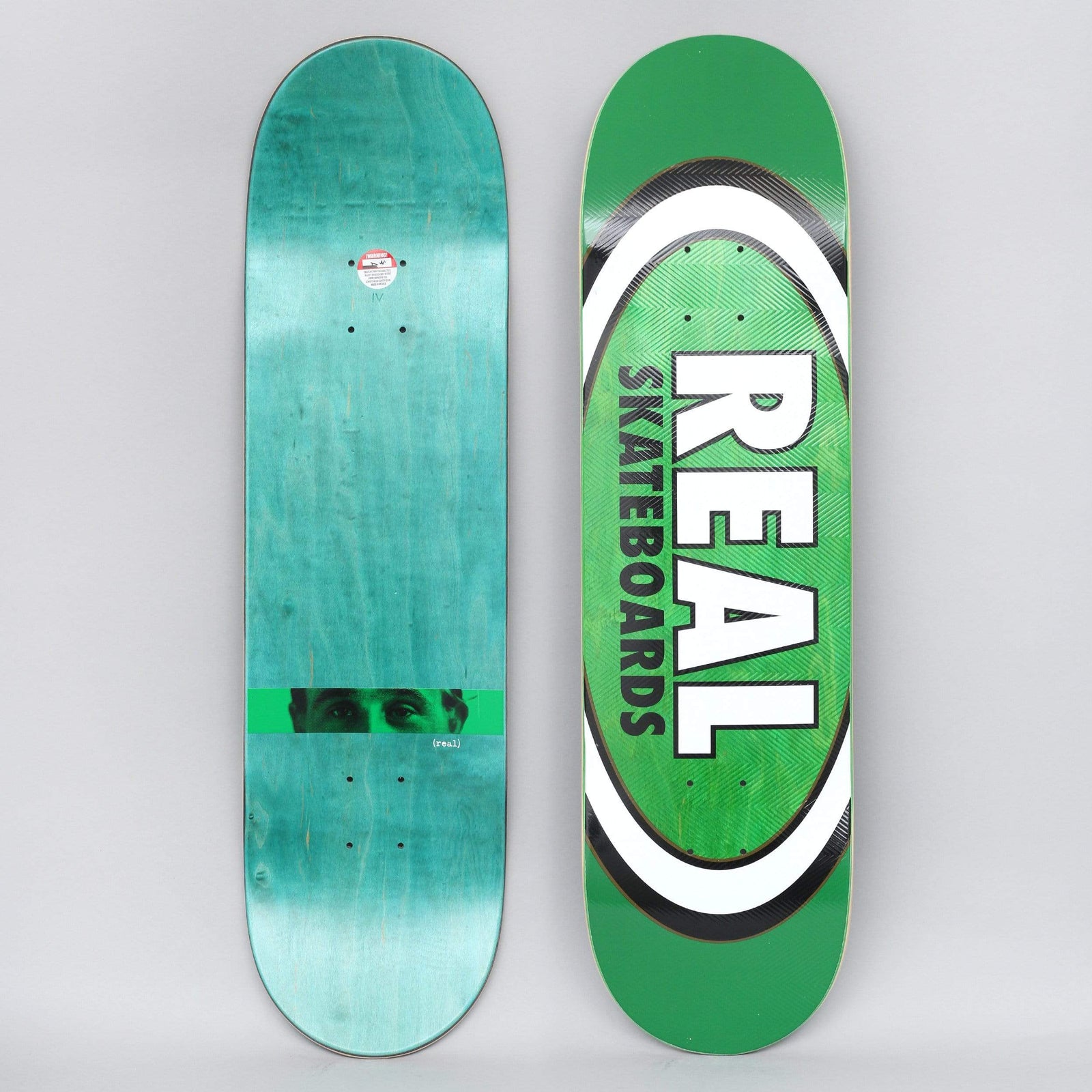 Real Harry Am Edition Oval Green skateboard deck: Large Real Skateboards oval logo in white and black over a green deck, with white capitals reading REAL SKATEBOARDS. The underside shows a close-up crop of eyes across a green band on teal wood stain.