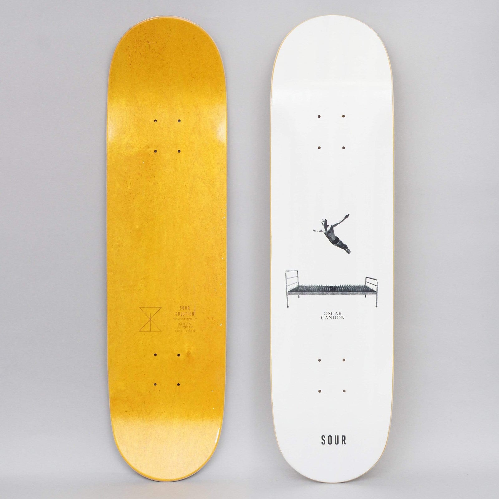 Sour Oscar Bed Of Nails White skateboard deck: Minimal white deck with a small photo cut-out of a figure diving through the air above a metal bed frame with a bed of nails, with a name and the SOUR wordmark below. The underside is yellow-stained.
