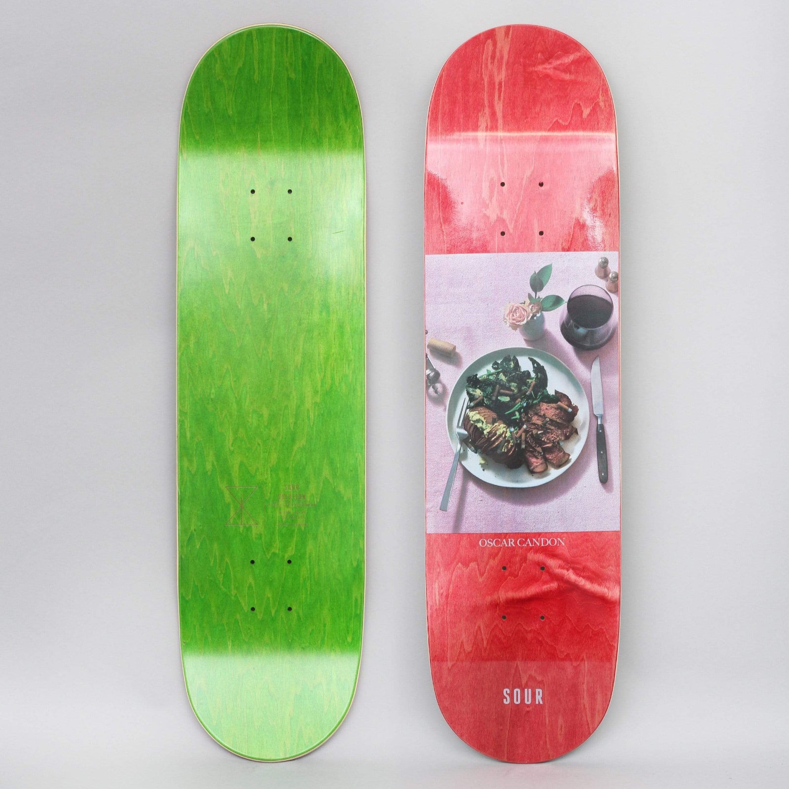 Sour Oscar Gourmet Red skateboard deck: A photographic panel on a red-stained deck showing a plate of steak and greens on a pink tablecloth with a glass of red wine, a knife, a fork and a small rose. Rider name and brand logo printed below.