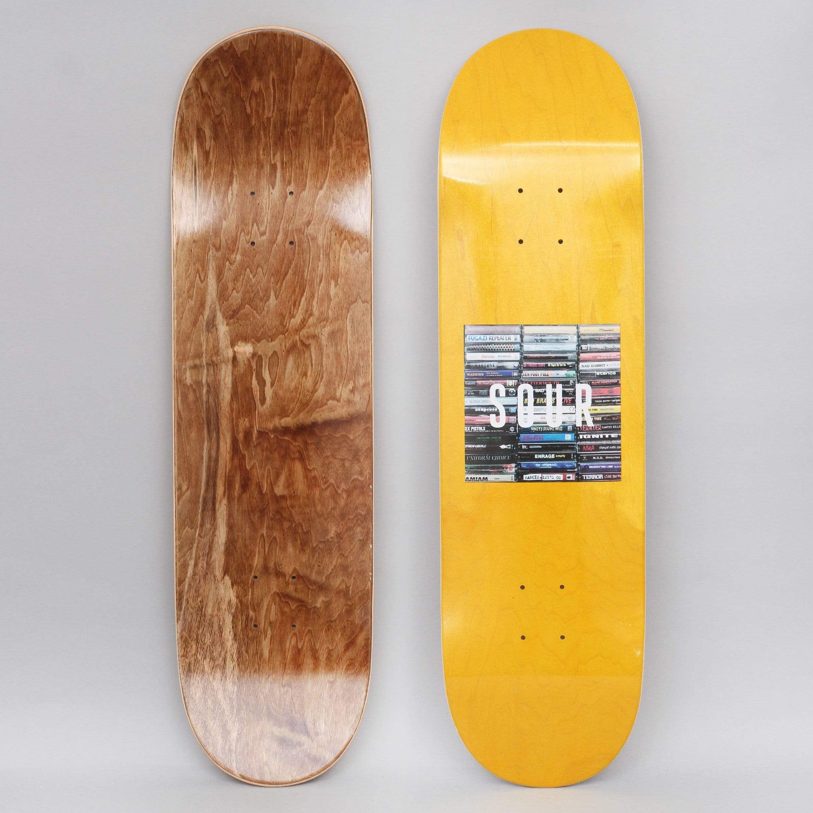 Sour Box Logo Tapes Yellow skateboard deck: Yellow-stained deck with a square photo of stacked cassette tapes and CDs and the white word SOUR across it. Brown wood underside shown alongside.