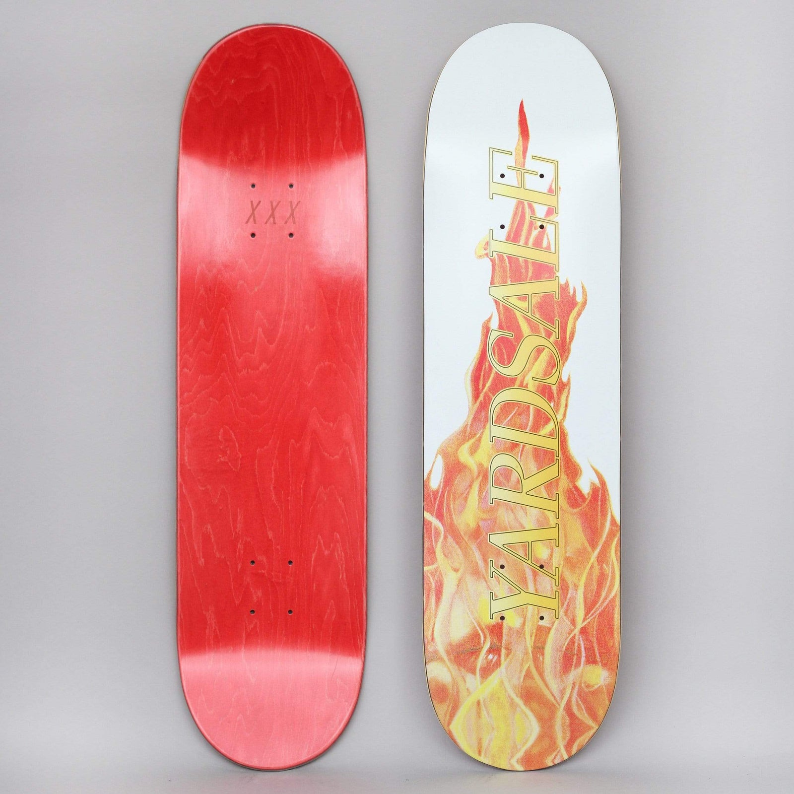 Yardsale Fuego White skateboard deck: Both sides shown. A photographic bonfire of orange, red and yellow flames rises up a white deck, with YARDSALE in gold serif capitals running vertically over it. The underside is red-stained with XXX near the top.