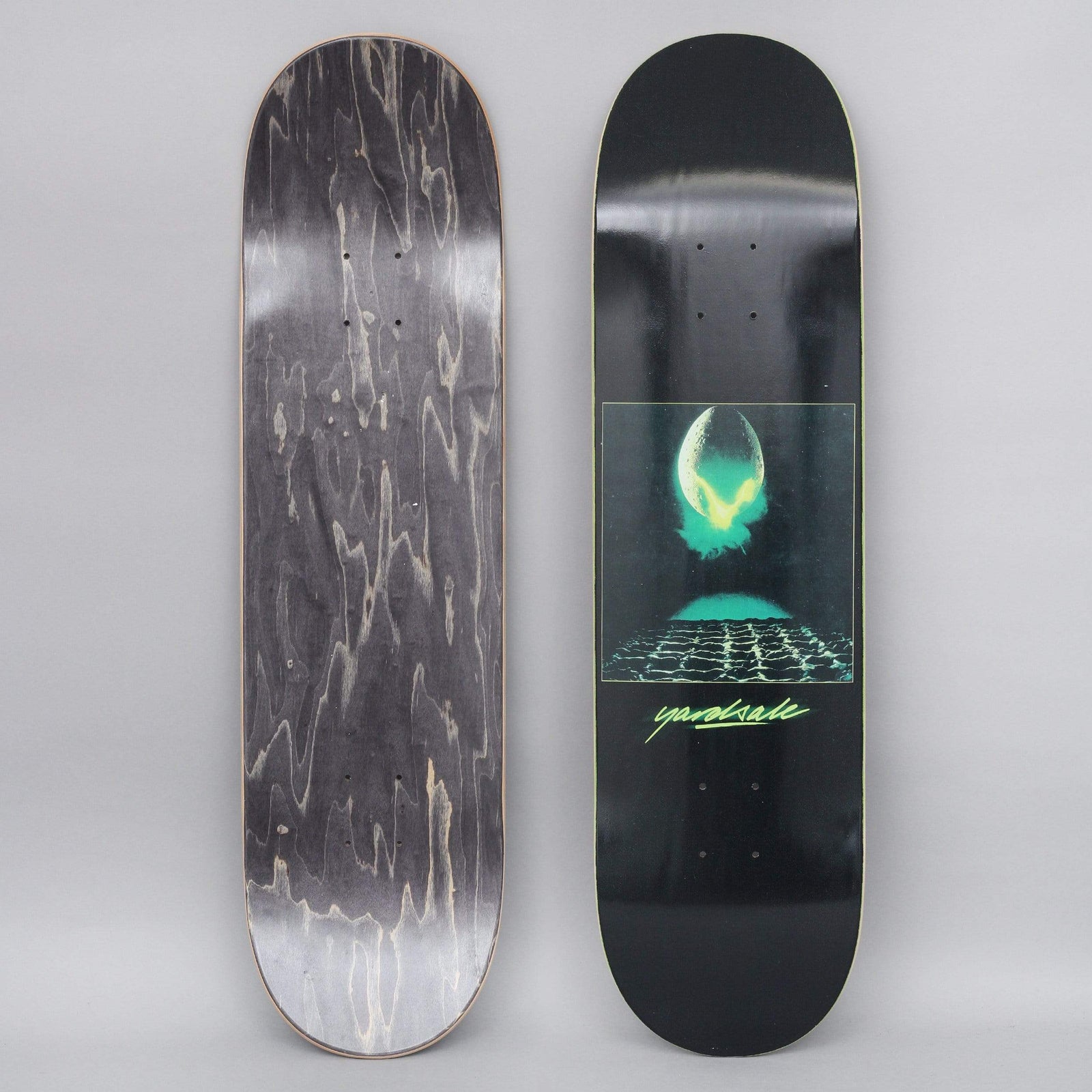 Yardsale Genesis Black skateboard deck: Black deck with a central framed image of a glowing green alien egg splitting open above a grid-patterned surface, in a sci-fi film-poster style. Green handwritten-style 'yardsale' logo underneath.