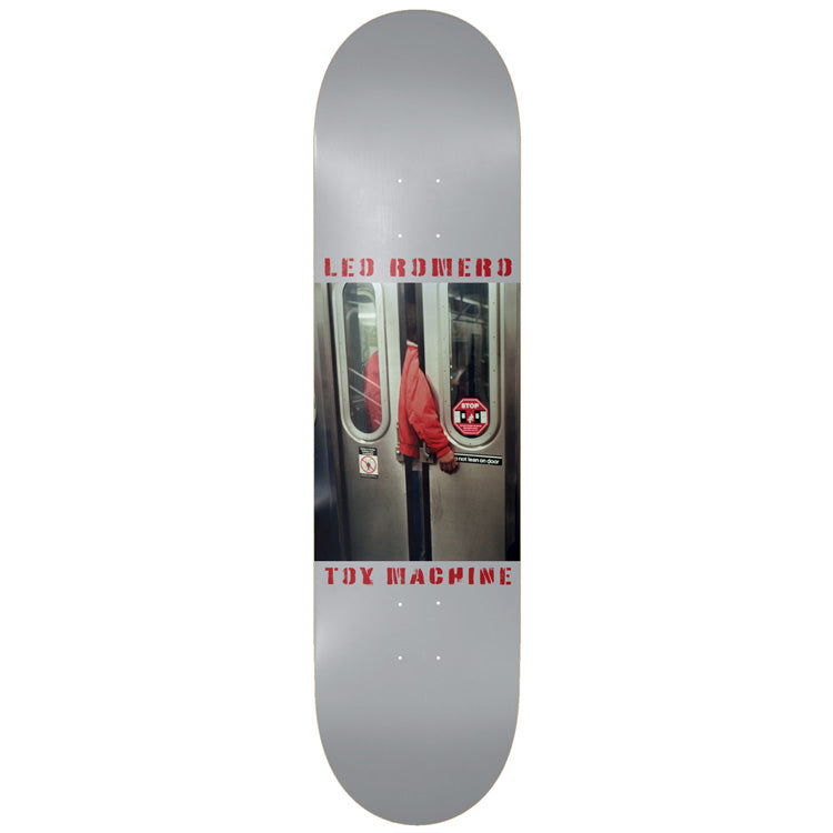 Toy Machine Leo Romero Wisecup 8.25 skateboard deck: Colour photograph of a person in a red jacket reaching an arm between closing subway train doors, set in a panel on a plain grey deck. Red stencil rider name above and Toy Machine below.