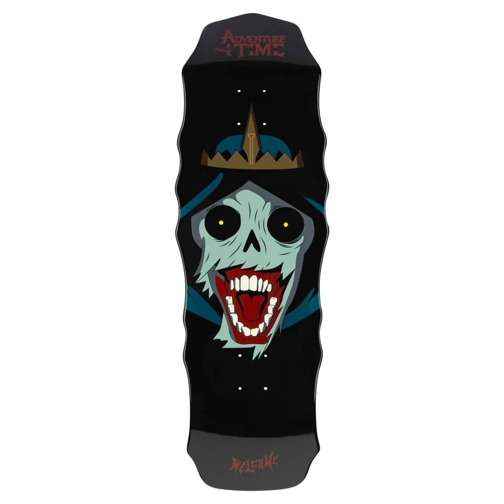 Welcome ADVENTURE TIME X WELCOME - The Lich on Widow skateboard deck: Screaming pale-green skull face with round black eyes and a red open mouth, wearing a hood and a gold spiked crown. Adventure Time logo at the nose, on a black wavy-edged shape.