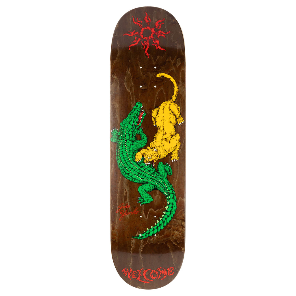 Welcome Jake Yanko Swamp on Popsicle - Brown Stain skateboard deck: A green alligator and a yellow panther fight, drawn in an engraved line style on a brown stained wood deck, with a red tribal sun at the top and red Welcome lettering and signature at the bottom.