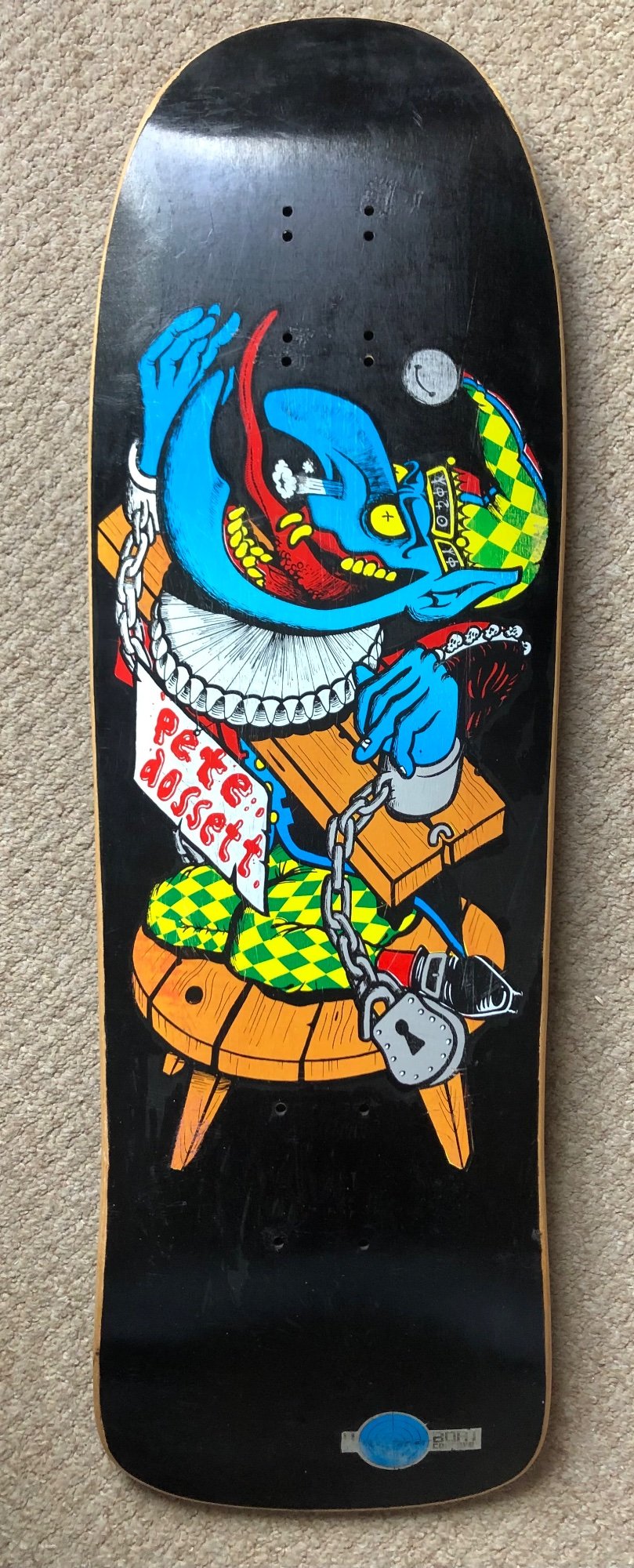 Deathbox Shackled (Different Colourway) skateboard deck: A blue-skinned jester in a ruff and green and yellow diamond-patterned costume sits on a wooden stool, wrists chained to a padlock, with a hand-lettered sign naming Pete Dossett. Bold cartoon outlines on a black background.