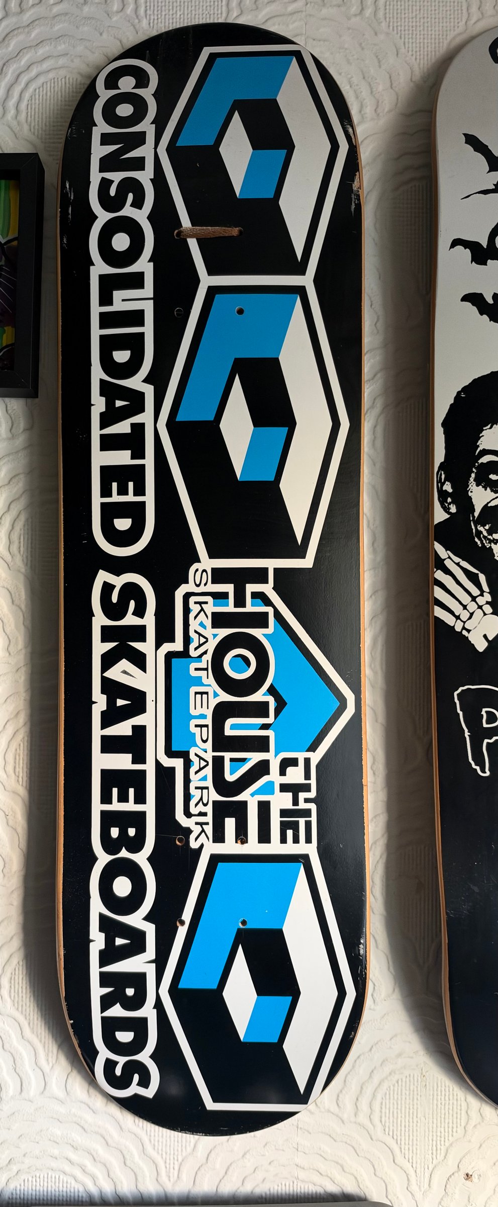 Consolidated House skatepark colab skateboard deck: Black deck with three white hexagonal cube emblems in blue and white running down its length, with Consolidated Skateboards lettered vertically up the side and a House Skatepark logo in the middle.