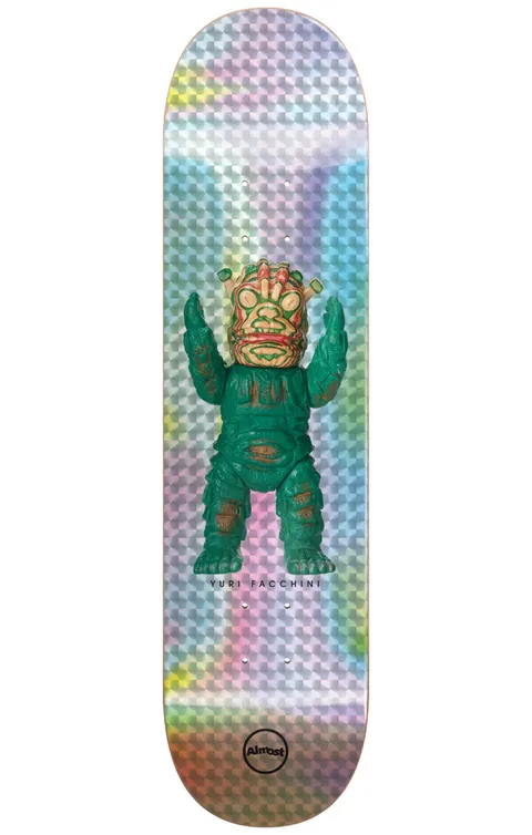 Almost Yuri Haroshi Monster Super Sap R7 8.125 & 8.375 skateboard deck: Holographic silvery deck with a prismatic square pattern, carrying a photographed green vinyl toy monster with a tiki-like mask face and raised arms. Yuri Facchini is printed beneath it, with the Almost circle logo at the tail.