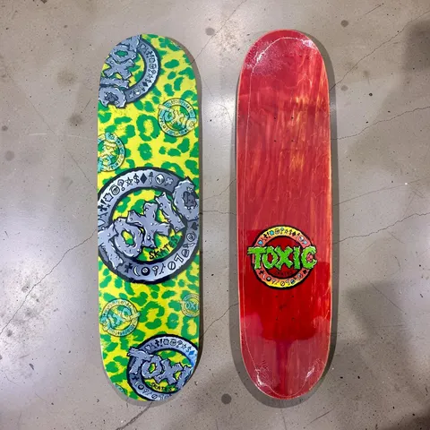 Toxic Toxic Team Leopard skateboard deck: Yellow deck with green leopard spots, overprinted with large grey chunky Toxic ring logos with studs and symbols, plus smaller teal ones. A red-stained underside shows a green Toxic logo in a circle.
