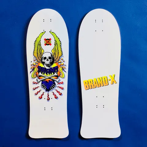 Brand-X X-Con Glow-in-dark skateboard deck: A retro skull and crossbones with yellow-green wings, entwined snakes and a heart with dripping red lines, with a purple 'Brand' scrawl across the skull. Old-school shaped white deck; the second deck shows a plain white back with a yellow-orange 'Brand-X' logo.