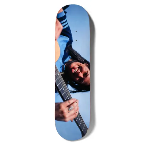 Girl Spike Photo Kim Deal skateboard deck: Colour photograph, rotated sideways, of a smiling dark-haired woman in a blue top playing an electric guitar against a blue sky; a ringed hand is on the fretboard.