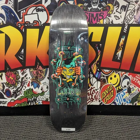 Black Label John Lucero X2 Tugboat skateboard deck: Orange-faced goblin jester in a green hat grips crossed bones within a studded cage frame, on a black wood-grain stain. Lucero is written in teal lettering below.