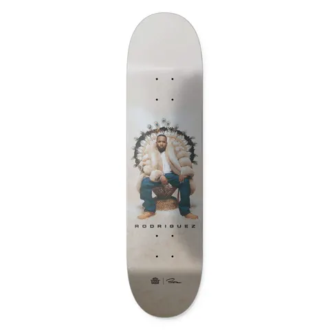 Primitive X Outkast Rodriguez Regal skateboard deck: Colour photograph of a man in a cream fur coat and jeans seated on a large wicker peacock chair edged with feathers, on a soft beige background. RODRIGUEZ in spaced capitals below, with Outkast and Primitive logos at the tail.