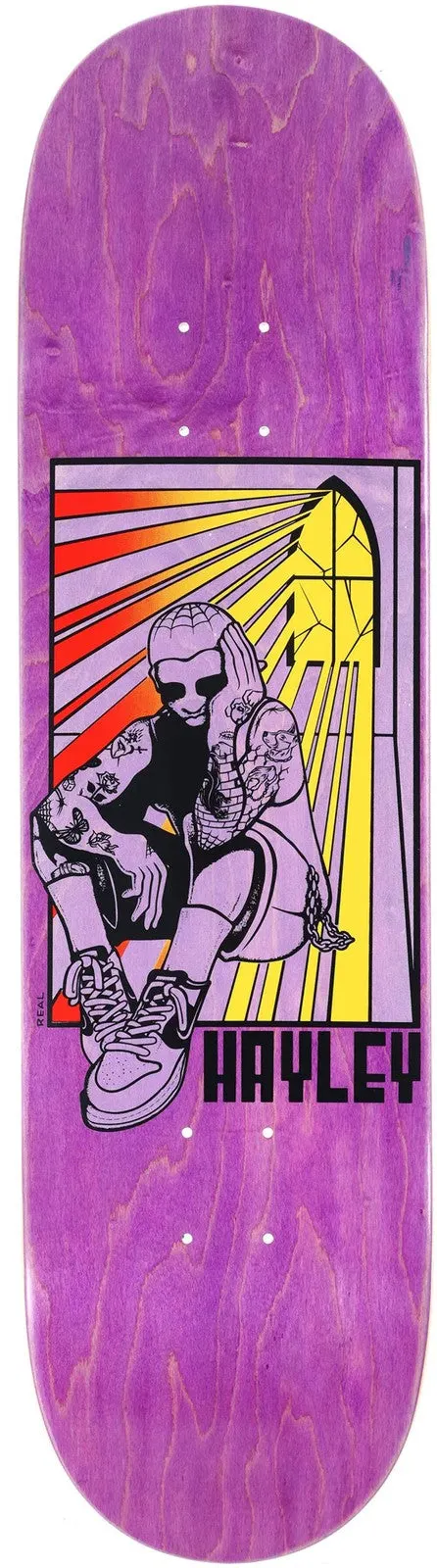 Real Hayley Stained skateboard deck: Stained-glass style panel on a purple wood-veneer deck showing a tattooed woman in sunglasses and high-top trainers sitting with her knee up, under rays of red, orange and yellow light from a window. The name is in block letters.