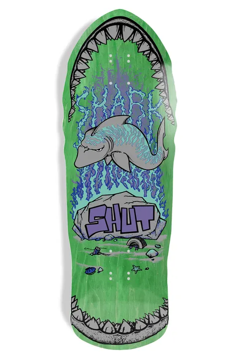 Shut 86 Shark EX7 (Green) skateboard deck: Old-school shaped deck on green stain showing a grey shark with a grumpy face swimming over a rock, with blue wavy water lines. The SHUT logo in purple graffiti letters sits on the rock, and shark jaws with teeth frame the nose and tail.