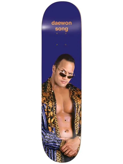 Enjoi Daewon Round 3 R7 8.25 & 8.5 skateboard deck: Photograph of a muscular man in tinted sunglasses and an open patterned gold and blue jacket, arms crossed low, on a deep blue deck with the rider name in orange at the nose.