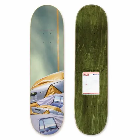 Habitat Paper Skaters De Keyzer 8.25 skateboard deck: A photographic image of cars covered in gold and silver protective sheeting under a hazy green-grey sky, with a thin gold vertical stripe. A postal-label style sticker is printed on the underside.