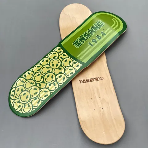 Insane Capsule Cruiser skateboard deck: Deck shaped like a green capsule pill, one half packed with yellow smiley faces and the other plain green with 'INSANE 1984' in dark outline lettering and light stripes. A natural wood deck with a small Insane logo lies behind.
