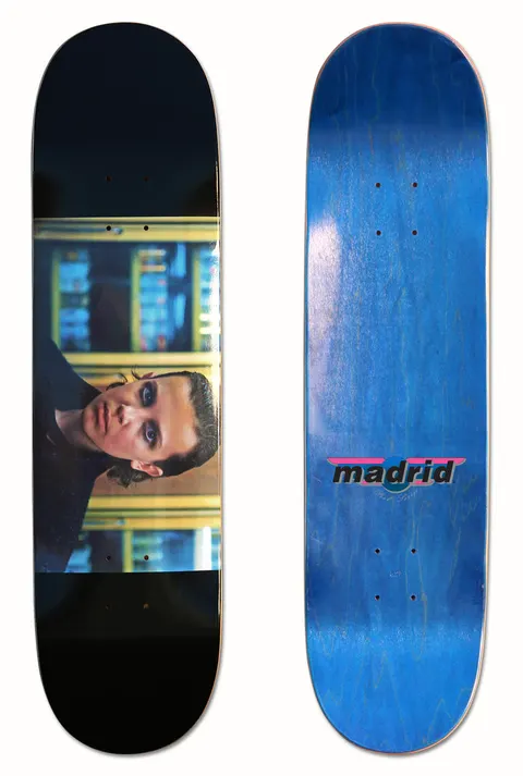 Madrid X Stranger Things - B*tchin Eleven 8 skateboard deck: Cinematic still of a young woman with short dark hair and heavy eye makeup, in dark clothes in front of shelves, as a wide horizontal photo band on a black deck. The blue underside carries the Madrid logo.