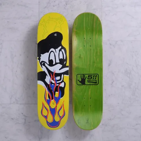Quasi Bigbiz Yellow skateboard deck: Both sides shown. A black-and-white cartoon duck-like character in a cap, grinning with blue and orange flames below its beak, on a yellow deck with faint green scribbles. The underside is green with a black hand logo.