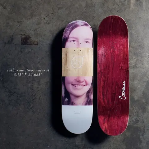 Quasi Catharine - Natural skateboard deck: Colour photographic portrait of a smiling young woman with long brown hair, split so a band of bare natural wood with a small white JJ line logo covers her nose and eyes' lower area. White tail. Red-stained underside shows Catharine in script.
