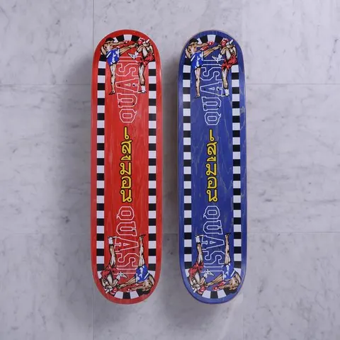 Quasi K.O. Red skateboard deck: Red-stained deck bordered with a black and white chequered edge, showing two Muay Thai fighters mid high kick at each end, with Quasi in outlined letters and yellow Thai script down the middle. Blue version shown beside it.