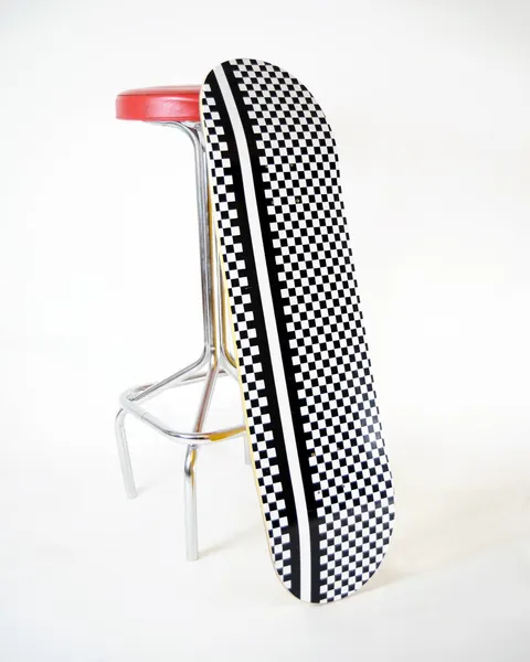Skateboard Cafe Check Stripe Black/White skateboard deck: Black and white chequerboard pattern covering the whole deck, with a solid white stripe and black stripe running down the length of it. Shown leaning against a bar stool.