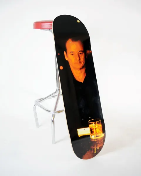 Skateboard Cafe LIT Bob skateboard deck: Dim, warm-lit photographic image of a man in dark clothing sitting at a bar, looking down at a glass of whisky in the foreground. Photo-style graphic covering the whole deck.