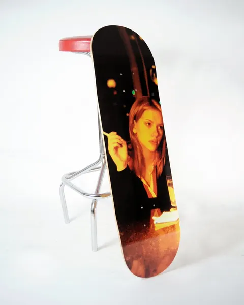 Skateboard Cafe LIT Charlotte skateboard deck: Warm-toned photograph of a woman with shoulder-length hair holding a cigarette, seated at a bar with a drink, with blurred amber lights behind her. Deck is photographed propped against a bar stool.