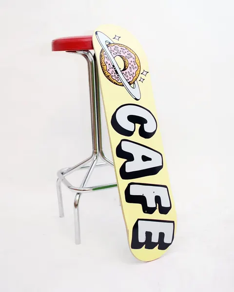 Skateboard Cafe Planet Donut Banana skateboard deck: Pink-iced sprinkled donut ringed like a planet, with sparkles, above chunky white Cafe lettering with black shadows on a pale yellow deck.