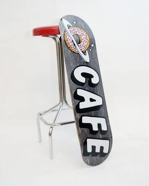Skateboard Cafe Planet Donut Black skateboard deck: Dark grey stained deck with a pink iced doughnut ringed like a planet with stars at the nose, above large white block letters spelling CAFE with black shadows.