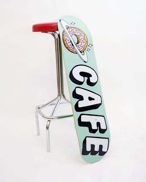 Skateboard Cafe Planet Donut Icing Blue skateboard deck: Pink-iced sprinkled doughnut ringed like a planet with sparkles above large white block letters reading CAFE with black drop shadows, on a pale mint deck.