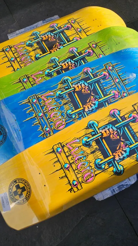 Black Label Lucero OG Bars Cross Shape (different colours) skateboard deck: Bars-style graphic of a cross of metal rods with studs and pink Lucero lettering, with a skull and hands in the centre, shown in yellow, green and blue stains. Stack of decks photographed at an angle.