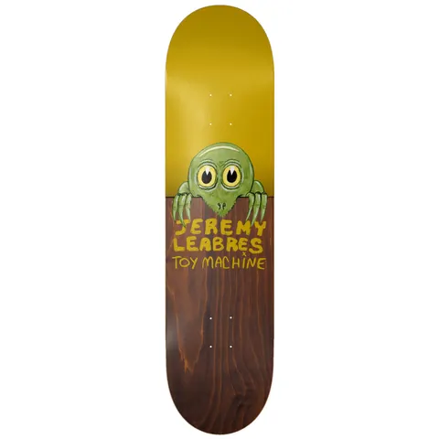 Toy Machine Jeremy Leabres Fence 8.50 skateboard deck: Cartoon green creature with big yellow eyes and long claws peering over a fence, formed by the split between a mustard-yellow upper deck and brown wood-stained lower half. Yellow hand-lettered names below.