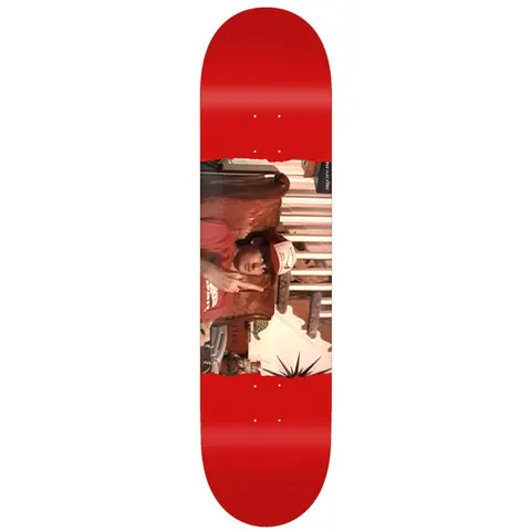 Toy Machine Ed Templeton Flip 8.50 skateboard deck: A rotated colour photograph of a person in a red top and cap, sitting in an ornate chair with a hand raised, printed as a band across a plain red deck. A small spiky starburst sits at the lower edge.
