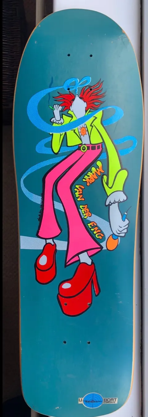 Deathbox Mini Van Der Eng (Different Colourway) skateboard deck: A cartoon figure with spiky red hair, a lime-green jacket, flared pink trousers and huge red platform shoes, smoking with blue swirling smoke curling around the body, on a teal background. The rider's name is written diagonally in orange lettering.