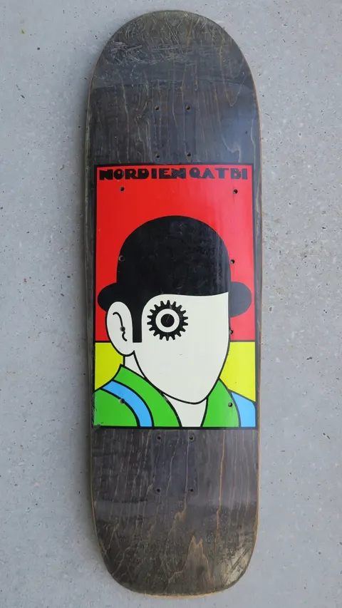 Deathbox Nordien Qatbi skateboard deck: Bold graphic panel of a faceless figure in a black bowler hat with a gear-like eye, green jacket with blue stripe, on red, yellow and green bands. Name lettering sits across the top, on a black-stained cruiser-shaped deck.